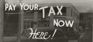 Writing on a window saying, "Your tax now here!" A building and a car are relfected in the window.