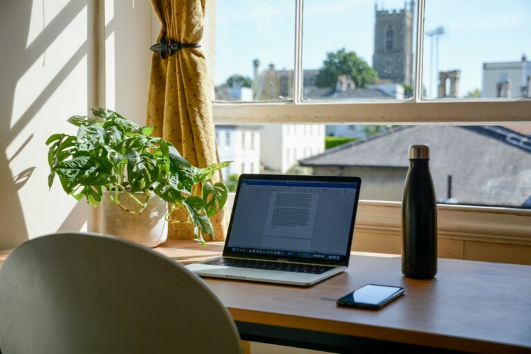 Laptop, mobile phone, water bottle and houseplant on brown wooden table. Open sash window overlooks a church.