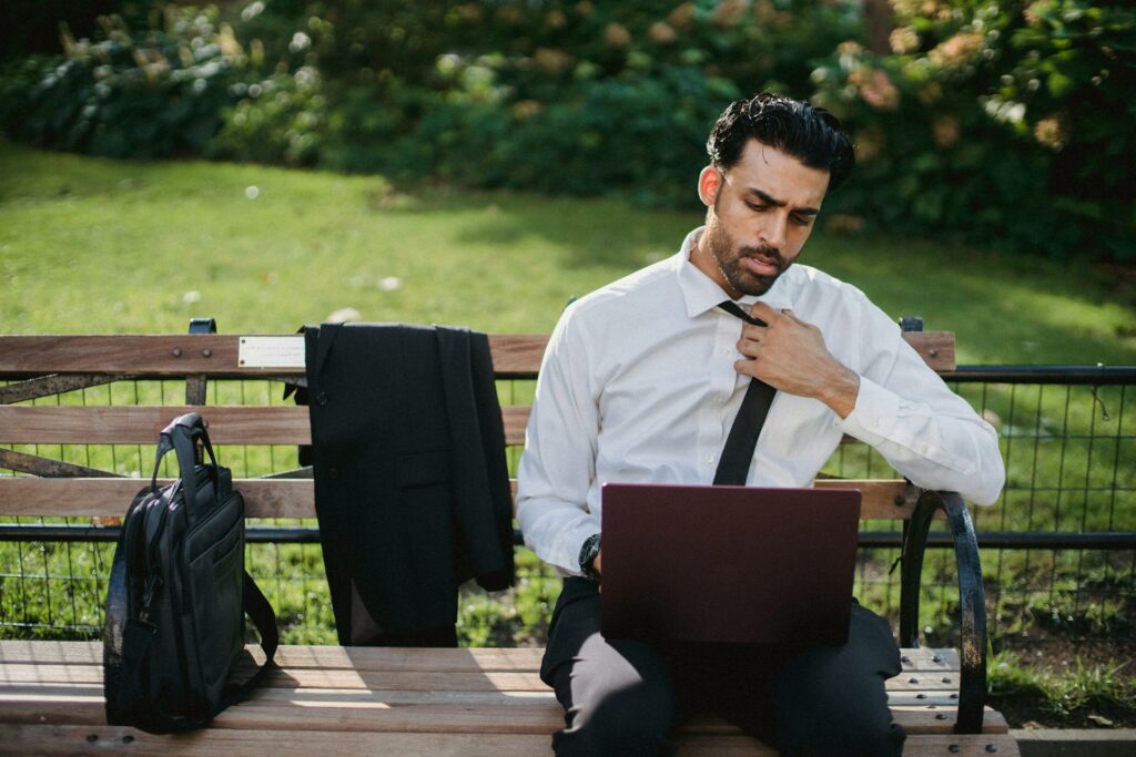 Man sitting on park bench with laptop on his lap. Suit jacket slung over the bench and briefcase on the bench.